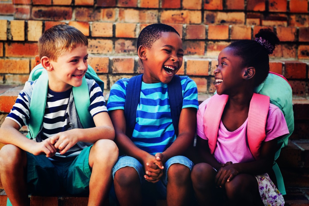 Three students with backpacks laughing together outside a brick school building