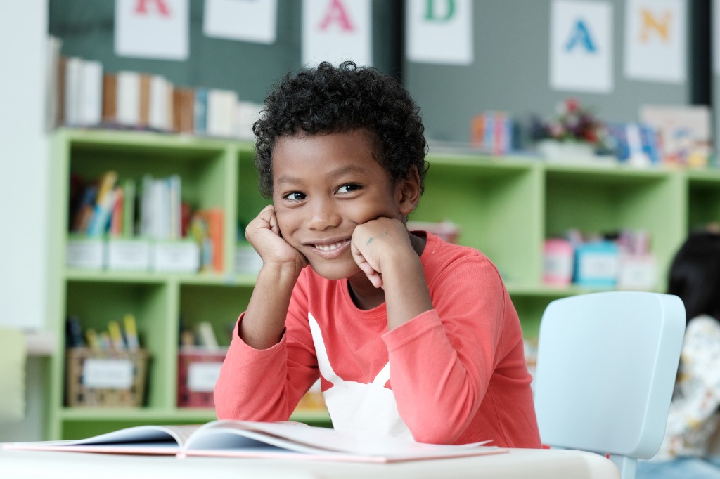 A young student smiling at a desk with an open book in a school library